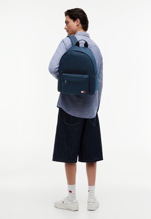 Kék inges és sötét rövidnadrágos személy tengerészkék hátizsákkal, fehér zoknival és fehér tornacipővel egy egyszerű fehér háttér előtt.