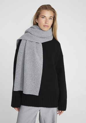 Écharpe grise côtelée en matériau doux, drapée autour d'un pull noir en maille épaisse avec un ourlet large et côtelé. Le gris clair contraste avec le ton foncé.