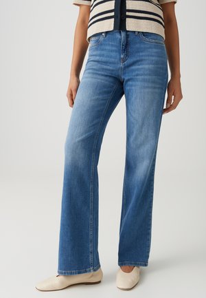 Frau trägt hellblaue ausgestellte Jeans, beige flache Schuhe und eine beige Strickjacke mit schwarzen Streifen, steht vor einem neutralen Hintergrund.