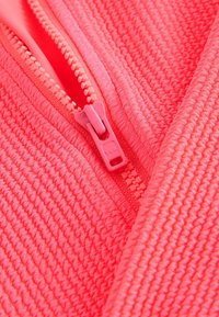 Helle pinke, mit Reißverschluss versehene Jacke aus einem strukturierten Stoff mit einem geriffelten Muster. Sichtbarer Kunststoffreißverschluss und Nähte.