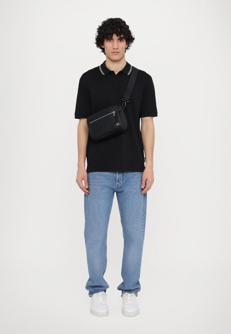 Polo nero con dettagli bianchi sul colletto, jeans in denim azzurro chiaro e sneakers bianche. È indossata una borsa a tracolla nera con chiusura a zip.