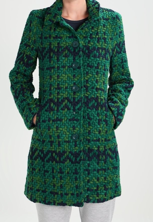 Manteau en laine vert et bleu foncé à motifs, avec de gros boutons et des poches latérales, porté sur un pantalon gris clair et un haut noir.