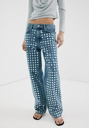 Persona vistiendo jeans azules de talle alto con tachuelas circulares plateadas, combinados con un top gris de manga larga y corta, y sandalias negras de punta abierta.