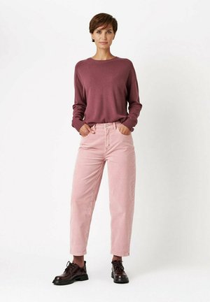 Woman standing with hands in pockets wearing a mauve sweater, light pink pants, brown socks, and dark brown lace-up shoes.