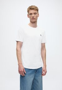 Bijela majica kratkih rukava od pamuka, s malim crnim logotipom na lijevoj prsnoj strani. U kombinaciji s kratkim hlačama od svijetloplog trapera.