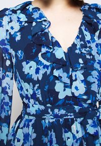 Robe fleurie bleu marine et bleu clair avec un col à volants et des manches longues. Elle présente une taille ceinturée et un tissu fluide et texturé.