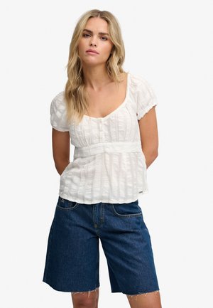 Frau mit langen blonden Haaren, die eine weiße, strukturierte kurzärmelige Bluse und dunkle Jeansshorts bis zum Knie trägt, steht mit den Händen hinter dem Rücken.
