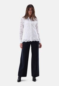 White lace blouse with long sleeves, button front, floral pattern, and scalloped hem, paired with wide-leg navy trousers and black ankle boots.