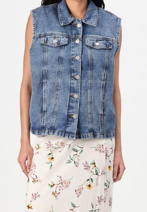 Mouwloos blauw denimvestje met knopen aan de voorkant en klepzakken, gedragen over een witte rok met groene en gele bloemenprint.
