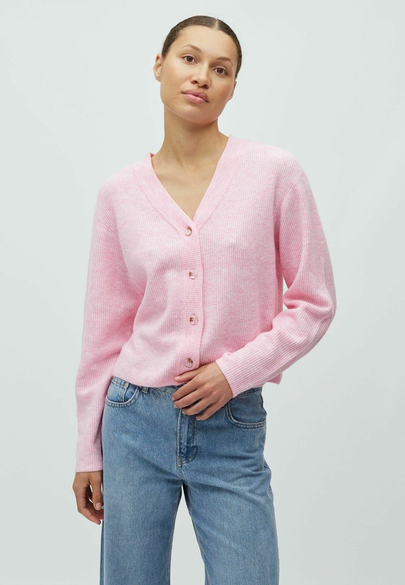 Roze geribde cardigan met V-hals en vijf knopen. Lange mouwen, zachte textuur, gecombineerd met een blauwe spijkerbroek, op een neutrale achtergrond.