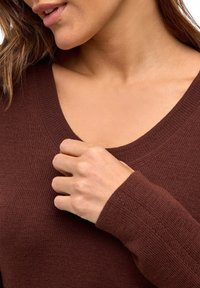 Eine Frau, die mit ihrer linken Hand den Ausschnitt eines braunen V-Ausschnitt-Strickpullovers in der Nähe des Schlüsselbeinbereichs anpasst.