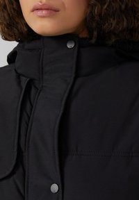 Crna puf jakna s visokim ovratnikom, s kopčanjem na dugmiće i vidljivim šavovima na prednjoj strani. Glatka tekstura tkanine.