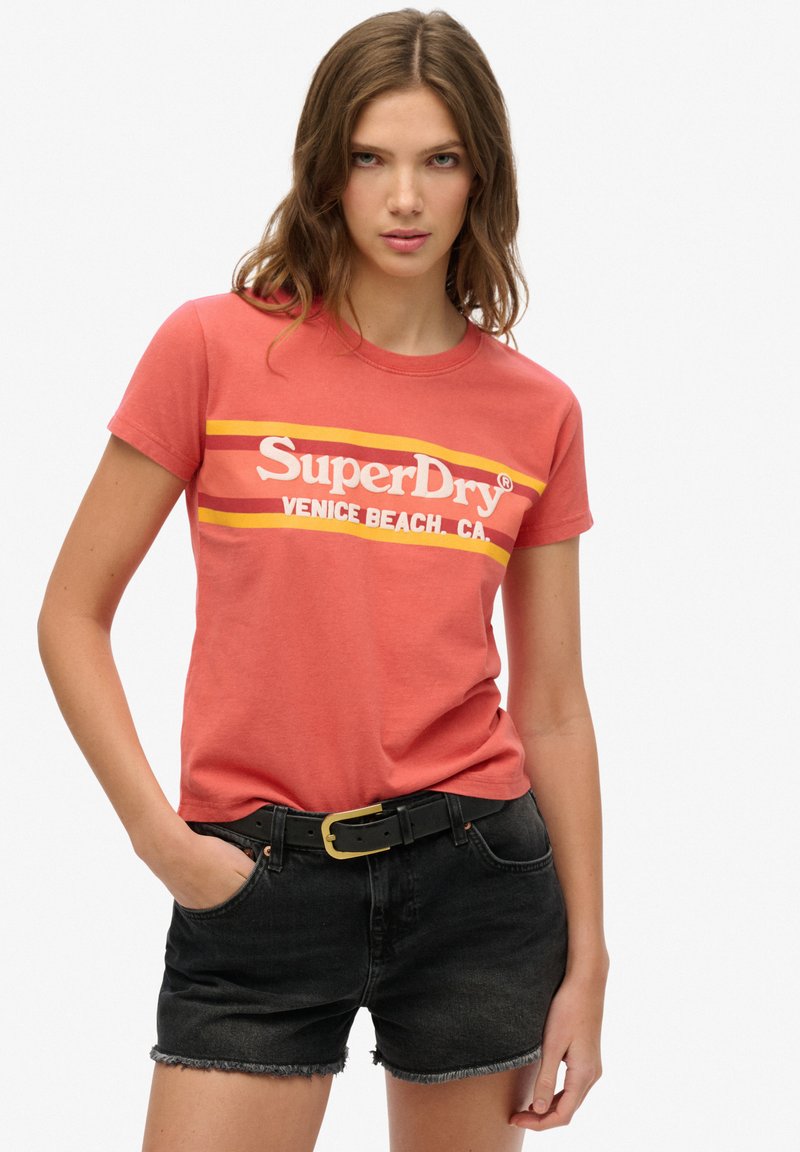 Kvinde stående med den ene hånd i lommen iført en rød SuperDry Venice Beach t-shirt og sorte denimshorts med sort bælte.