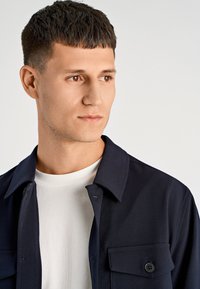 Marineblauw overhemd met knopen en twee borstzakken, gemaakt van gladde stof; gedragen over een wit T-shirt met ronde hals. Strak ontwerp met een moderne pasvorm.