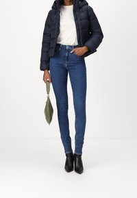 Donna con giacca imbottita scura, t-shirt bianca Calvin Klein, jeans skinny blu, stivaletti neri alla caviglia, che tiene una borsa verde per la tracolla.