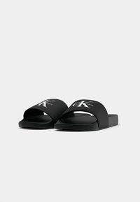 Black Calvin Klein slide sandals with wide straps and white logo text, positioned side by side on a plain light background.