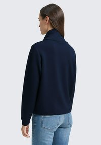 Dunkelblaues Sweatshirt mit hohem Kragen, strukturiertem Stoff und gerippten Bündchen, kombiniert mit hellblauen, schmal geschnittenen Jeans.