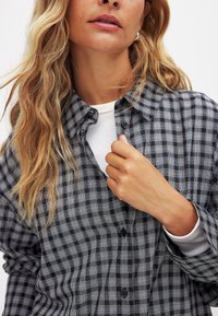 Black and white checkered button-up shirt with a textured, woven pattern, featuring a collared neckline and single button placket.