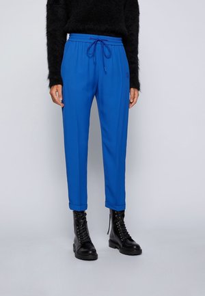 Person iført lyseblå snøre-bukser, sort fuzzy sweater og sorte snørestøvler står på en lys baggrund.