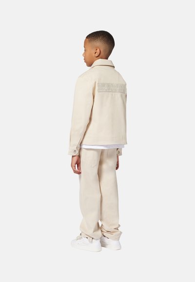 Beige jasje met een gestructureerd achterpaneel, bijpassende broek en witte sneakers, met een relaxte pasvorm en klassiek kraagontwerp.