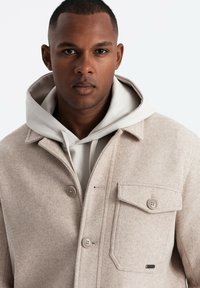 Veste en mélange de laine beige avec un bouton à l'avant, deux poches sur la poitrine et un sweat à capuche gris en dessous. Texture lisse, design classique.
