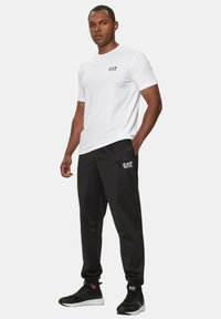 T-shirt atletico bianco con logo EA7 nero, abbinato a pantaloni della tuta neri con logo corrispondente. Scarpe da ginnastica nere completano l'outfit.