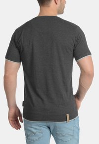 Graues Kurzarm-T-Shirt mit kontrastierendem hellgrauen Besatz an den Ärmeln. Etikett ist an der unteren Seite sichtbar, getragen mit hellblauen Jeans.