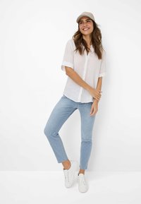 Hvid kortærmet skjorte med knaplukning, lyseblå stribede jeans og hvide sneakers. Modellen bærer en beige cap med lyseblå akcenter.