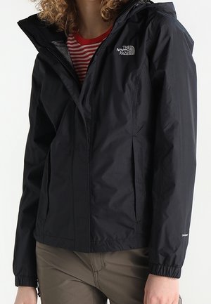 Black North Face waterproof jacket with hood worn over red and white striped shirt, paired with olive green pants.
