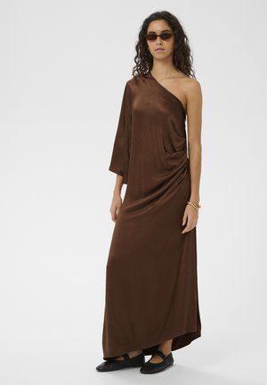 Donna che indossa un abito lungo marrone a una spalla, scarpe basse nere, occhiali da sole e braccialetto d'oro, in piedi contro uno sfondo chiaro e uniforme.