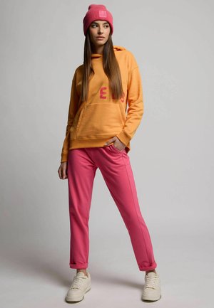 Orange hoodie with front pocket and pink text, paired with pink sweatpants and white sneakers. Pink knitted beanie on the head.