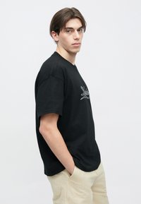 Νεαρός άνδρας με μαύρο μπλουζάκι με γραφικό κείμενο, μπεζ παντελόνι, στεκεται πλάγια με τα χέρια στις τσέπες, ουδέτερη έκφραση, απλό φωτεινό φόντο.