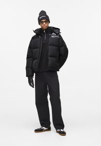 Zwarte pufferjack met een glanzend ontwerp en zilveren accenten, gecombineerd met een zwarte turtleneck, zwarte broek en zwarte sneakers met witte details.