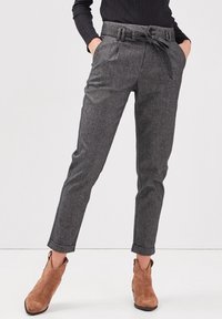 Kvinna iklädd grå skräddarsydda byxor med hög midja och knuten bälte, uppvikt fåll, svart topp och bruna mocka ankelskor. Händerna i fickorna.