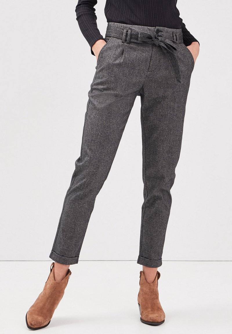 Kvinna iklädd grå skräddarsydda byxor med hög midja och knuten bälte, uppvikt fåll, svart topp och bruna mocka ankelskor. Händerna i fickorna.