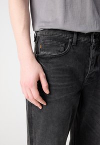 Jeans de mezclilla negra con un ajuste slim, que presentan un desvanecimiento mínimo, un bolsillo frontal con una etiqueta de logo naranja y una textura suave.