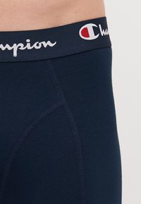 Mornarice bokserice s mekim elastičnim pojasom. Sadrže bijeli "Champion" logo i kontrastne šavove na tkanini. Glatka tekstura.