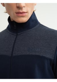 Close-up van een persoon die een marineblauw- en grijs ritssluitingjack met een hoge kraag en het logo "BIG STAR" op de borst draagt.