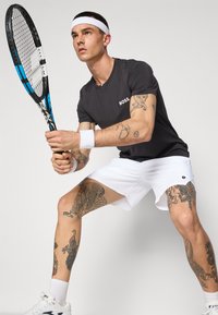 Svart treningstrøye med "BOSS"-logo, hvite shorts og et hvitt hodebånd. Holder en blå og svart tennisracket, og viser detaljerte tatoveringer.