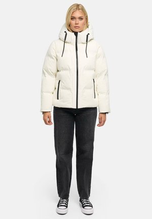 Femme portant une doudoune blanche avec capuche, un jean noir et des baskets noires, debout face à l'avant sur un fond uni.