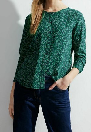 Femme portant un chemisier vert à motifs boutonné à manches longues et un jean bleu foncé, avec une main dans sa poche.