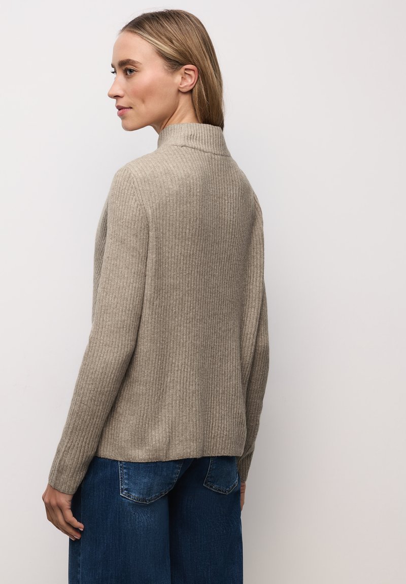 Pull côtelé beige avec un col montant et des manches longues, vu de dos, associé à un jean bleu à jambes larges. Texture lisse.