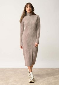 Abito lungo in maglia a trecce color taupe chiaro con scollo alto e silhouette aderente; presenta polsini e orlo a coste. Indossato con sneaker e calze.