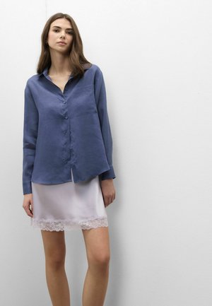Jonge vrouw die een losse blauwe blouse met knopen draagt en een witte rok met kanten randje, staand tegen een effen witte muur.