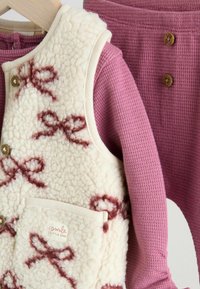 Fleece-Weste mit cremefarbigem Grund und burgunderfarbenem Schleifenmuster, kombiniert mit einem strukturierten rosa Langarmshirt und passenden Hosen mit Knöpfen.