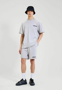 Joven con camiseta de ellesse gris claro y pantalones cortos, sombrero tipo cubo negro, calcetines blancos y zapatillas negras, de pie con una mano en el bolsillo.
