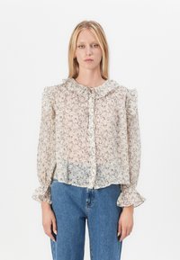 Blouse transparente avec imprimé floral, col et poignets volantés, fermeture à boutons et texture légère, associée à un jean en denim taille haute.