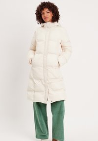 Manteau long matelassé en crème avec capuche, doté d'une fermeture éclair à l'avant et de deux poches latérales ; porté sur un pantalon en velours côtelé vert.