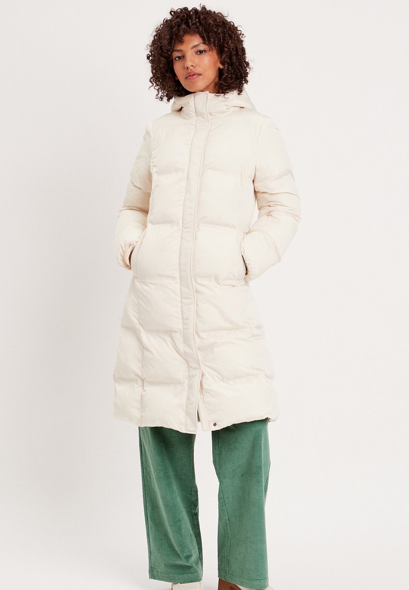 Manteau long matelassé en crème avec capuche, doté d'une fermeture éclair à l'avant et de deux poches latérales ; porté sur un pantalon en velours côtelé vert.