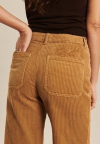 Pantalons en velours côtelé de couleur moutarde avec une texture rayée verticale, dotés de deux poches arrière et d'un design à coupe décontractée.